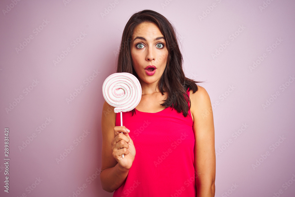 Young beautiful woman eating sweet candy over pink isolated background scared in shock with a surprise face, afraid and excited with fear expression Young beautiful woman eating sweet candy over pink isolated background scared in shock with a surprise face, afraid and excited with fear expression