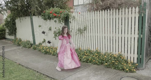 A little girl dressed in a princess costume dances, spins and twirls outside her house in front of a fence and flowers before curtsying and running off photo