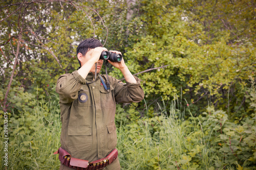 A male hunter with binoculars looks out for his prey against the background of the forest. The concept of a hunter, successful or failed hunt. Image.
