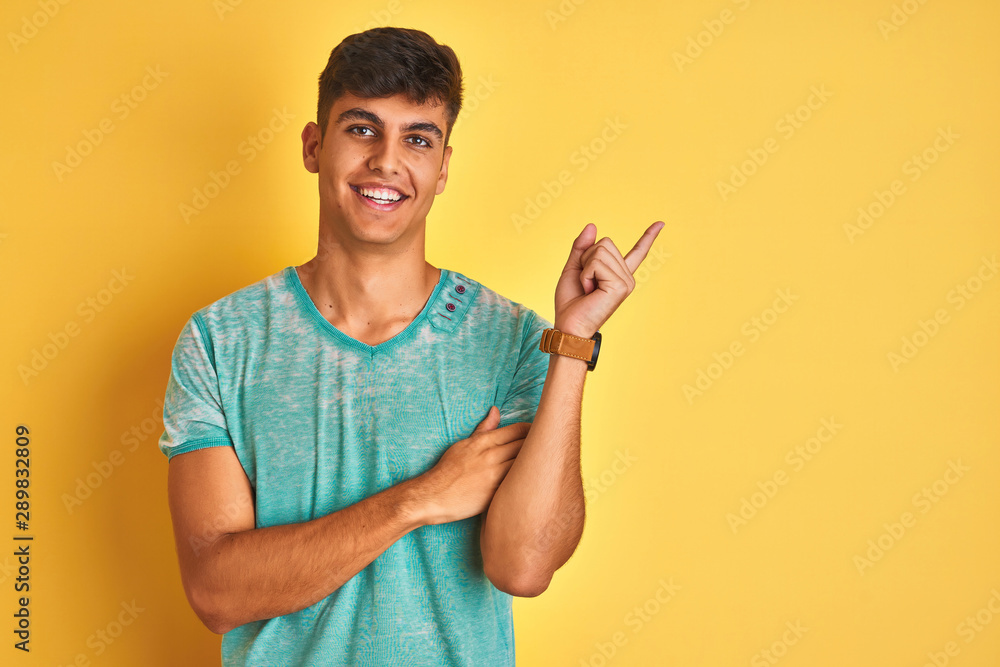 Young indian man wearing green t-shirt standing over isolated yellow background with a big smile on face, pointing with hand and finger to the side looking at the camera. Young indian man wearing green t-shirt standing over isolated yellow background with a big smile on face, pointing with hand and finger to the side looking at the camera.