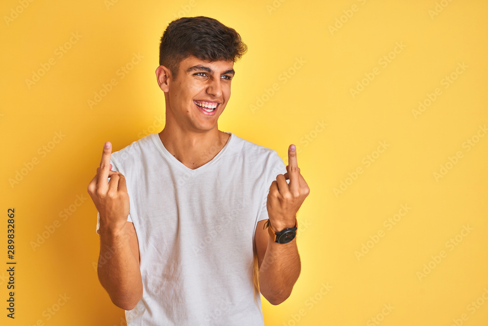 Young indian man wearing white t-shirt standing over isolated yellow background Showing middle finger doing fuck you bad expression, provocation and rude attitude. Screaming excited Young indian man wearing white t-shirt standing over isolated yellow background Showing middle finger doing fuck you bad expression, provocation and rude attitude. Screaming excited
