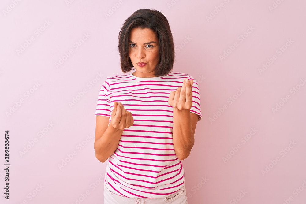 Young beautiful woman wearing striped t-shirt standing over isolated pink background doing money gesture with hands, asking for salary payment, millionaire business Young beautiful woman wearing striped t-shirt standing over isolated pink background doing money gesture with hands, asking for salary payment, millionaire business