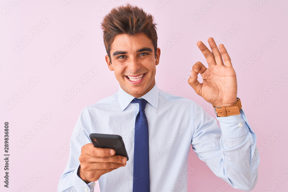 Young handsome businessman using smartphone standing over isolated pink background doing ok sign with fingers, excellent symbol Young handsome businessman using smartphone standing over isolated pink background doing ok sign with fingers, excellent symbol
