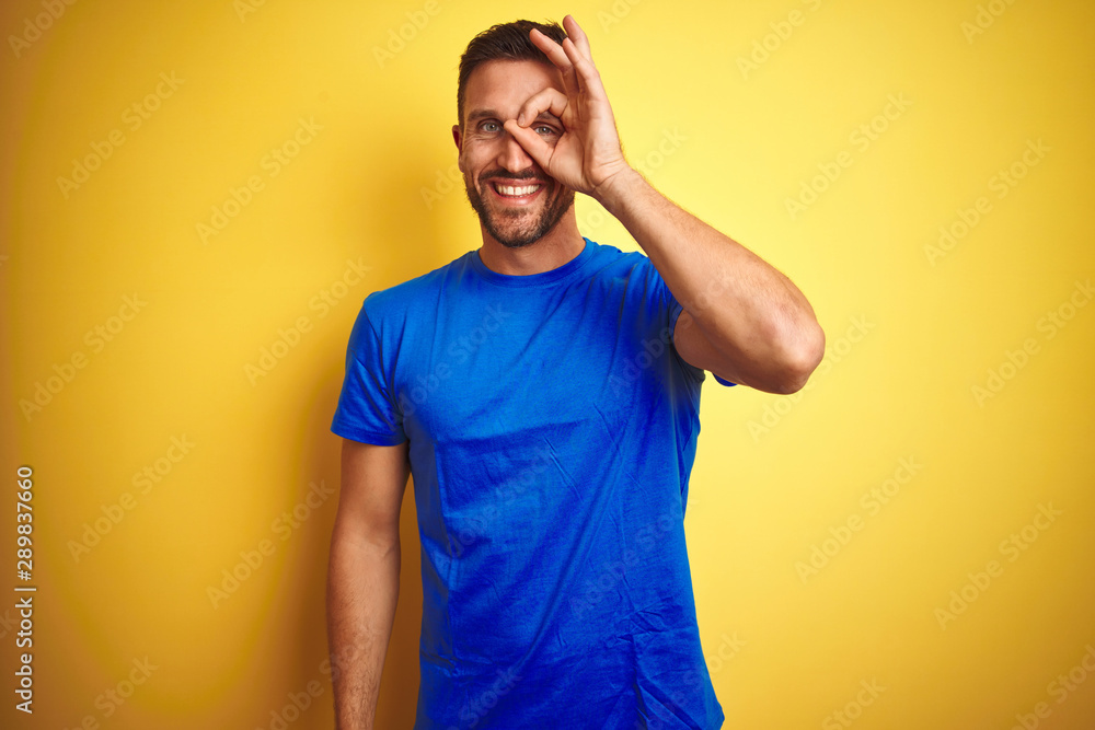 Young handsome man wearing casual blue t-shirt over yellow isolated background doing ok gesture with hand smiling, eye looking through fingers with happy face. Young handsome man wearing casual blue t-shirt over yellow isolated background doing ok gesture with hand smiling, eye looking through fingers with happy face.
