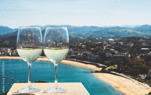 Two drink glass white wine stand on background blue sea top view city coast from observation deck, romantic toast with alcohol panoramic cityscape downtown, spain san sebastian vacation
