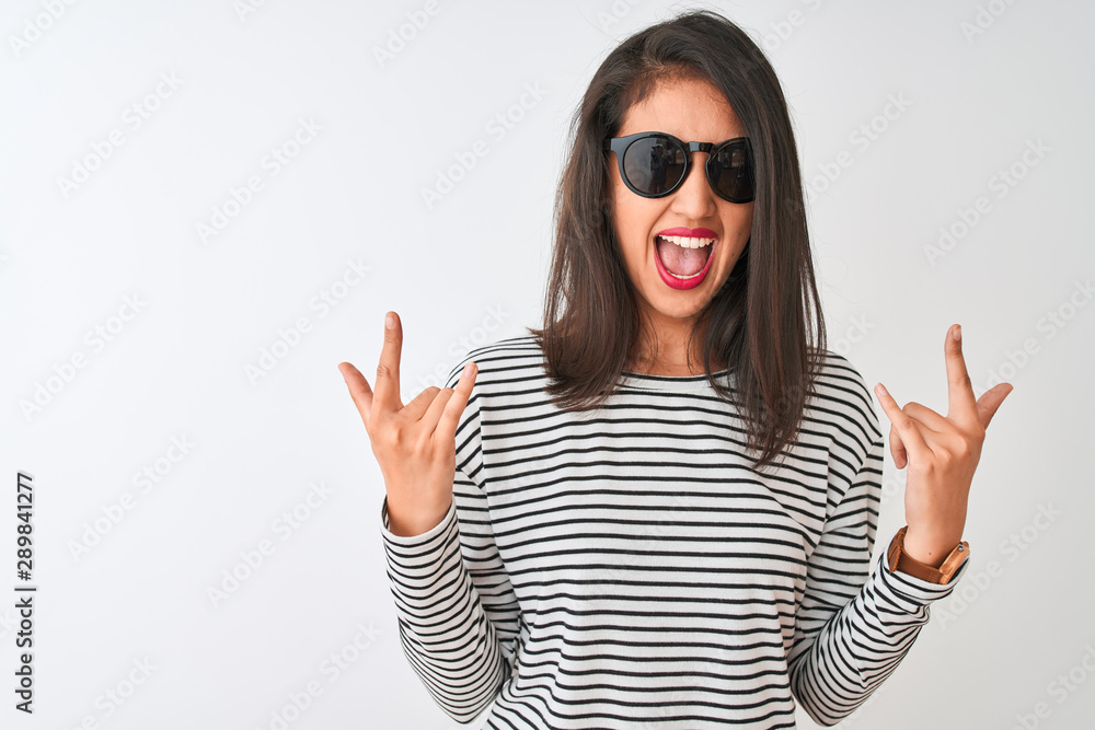 Chinese woman wearing striped t-shirt and sunglasses standing over isolated white background shouting with crazy expression doing rock symbol with hands up. Music star. Heavy concept. Chinese woman wearing striped t-shirt and sunglasses standing over isolated white background shouting with crazy expression doing rock symbol with hands up. Music star. Heavy concept.