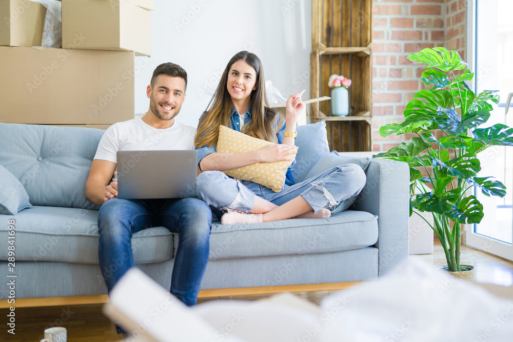 Young beautiful couple relaxing sitting on the sofa of new house using computer laptop very happy pointing with hand and finger to the side Young beautiful couple relaxing sitting on the sofa of new house using computer laptop very happy pointing with hand and finger to the side