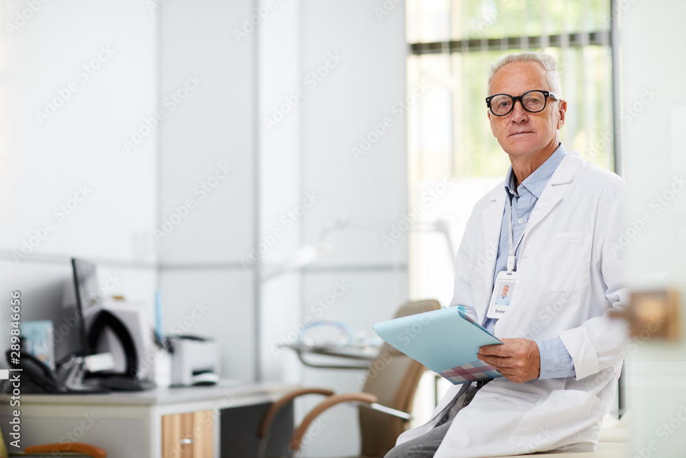 Portrait of professional senior doctor holding clipboard and looking at camera while taking notes standing in office of modern clinic, copy space