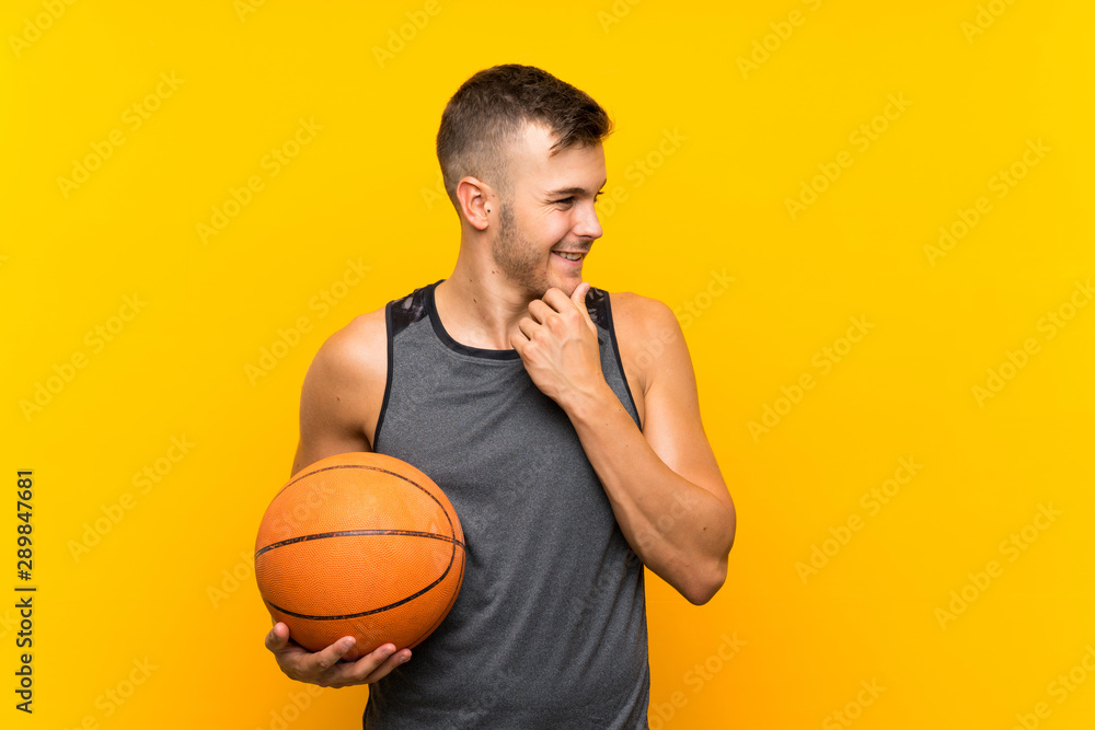 Young handsome blonde man holding a basket ball over isolated yellow background thinking an idea and looking side Young handsome blonde man holding a basket ball over isolated yellow background thinking an idea and looking side
