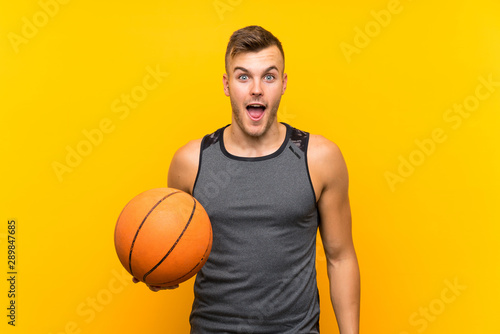 Young handsome blonde man holding a basket ball over isolated yellow background with surprise and shocked facial expression
