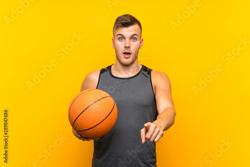 Young handsome blonde man holding a basket ball over isolated yellow background surprised and pointing front