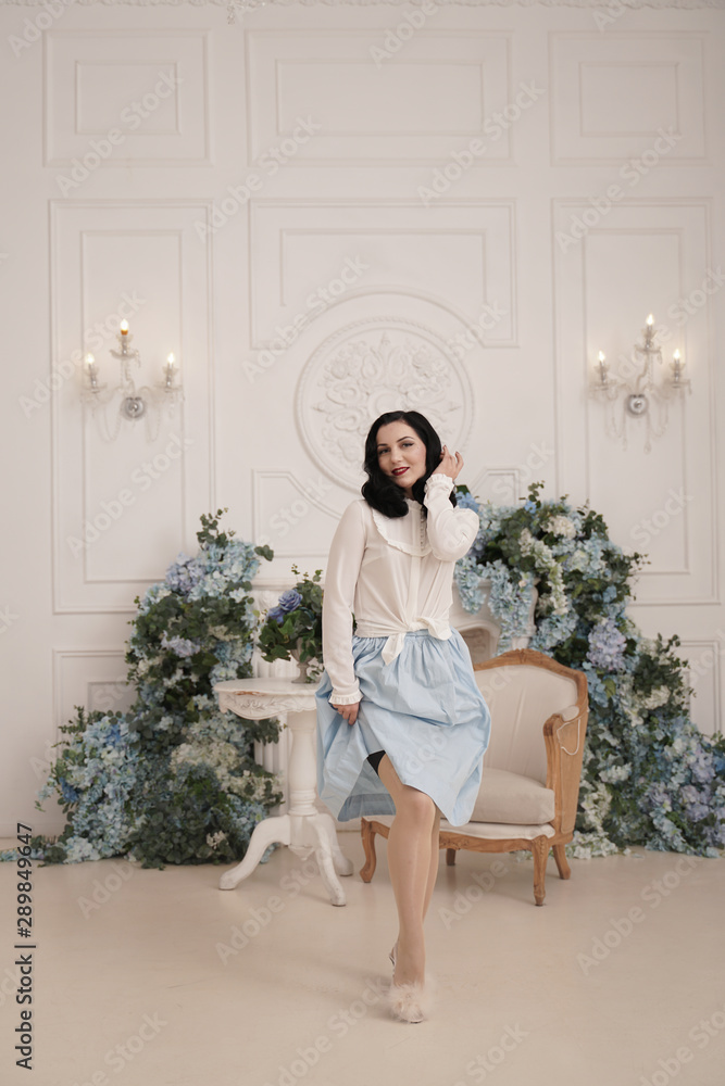 Lovely woman in retro cute cotton dress posing indoors in her white apartment. Fashion lady in vintage outfit with stockings. Lovely woman in retro cute cotton dress posing indoors in her white apartment. Fashion lady in vintage outfit with stockings.