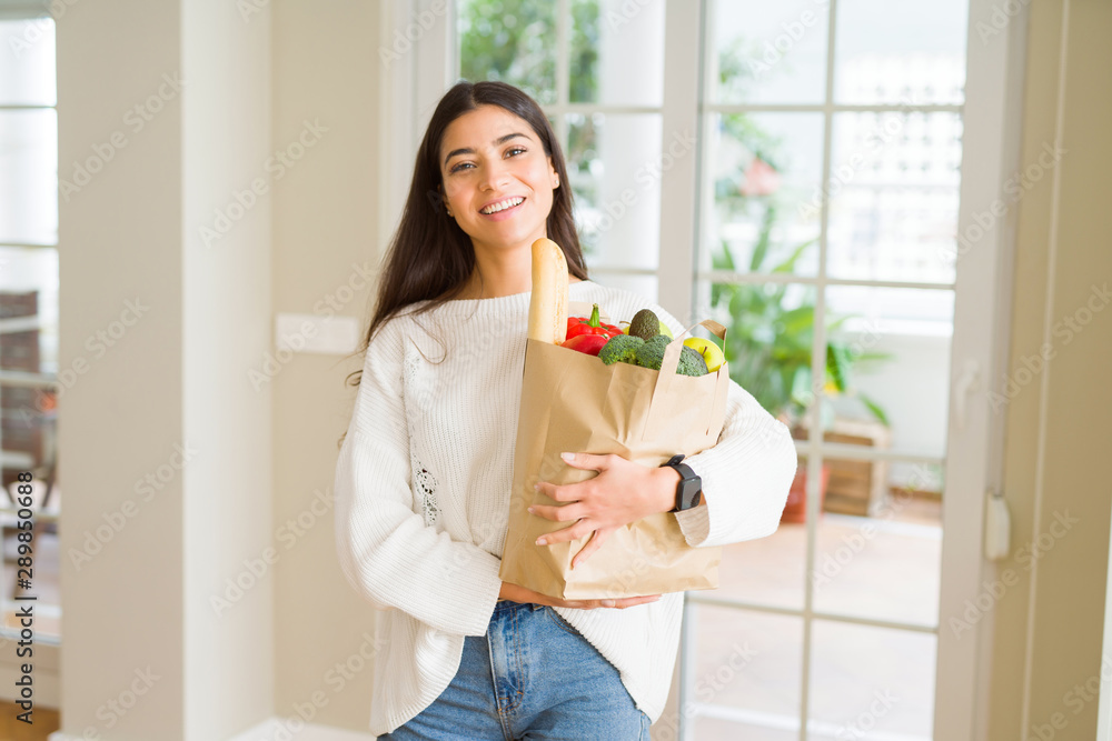 Beautiful young woman smiling holding a paper bag full of fresh groceries at home Beautiful young woman smiling holding a paper bag full of fresh groceries at home