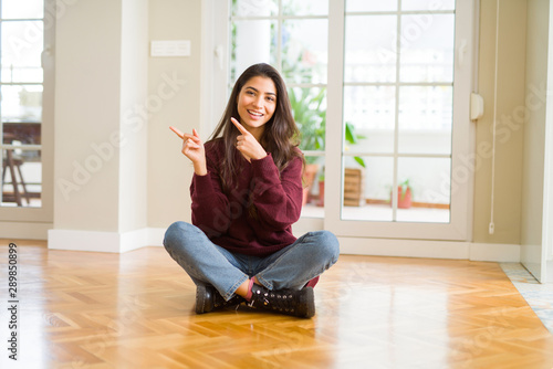 Free Download Young beautiful woman sitting on the floor at home smiling and looking at the camera pointing with two hands and fingers to the side. - GFXLoad