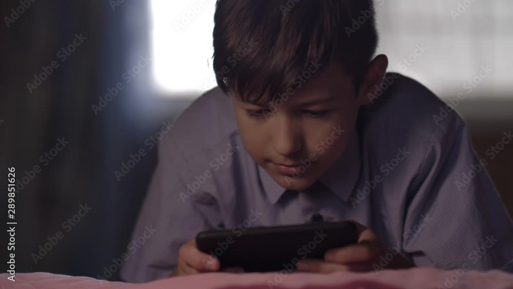 boy lies on the bed at home and uses a phone in the evening, plays games on the phone boy lies on the bed at home and uses a phone in the evening, plays games on the phone