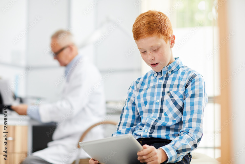 Portrait of cute red haired boy using digital tablet in waiting room at doctors office, copy space