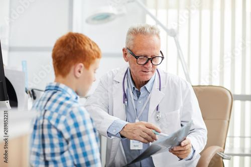Portrait of white-haired senior doctor pointing at x ray image while talking to little patient during consultation in child healthcare clinic, copy space