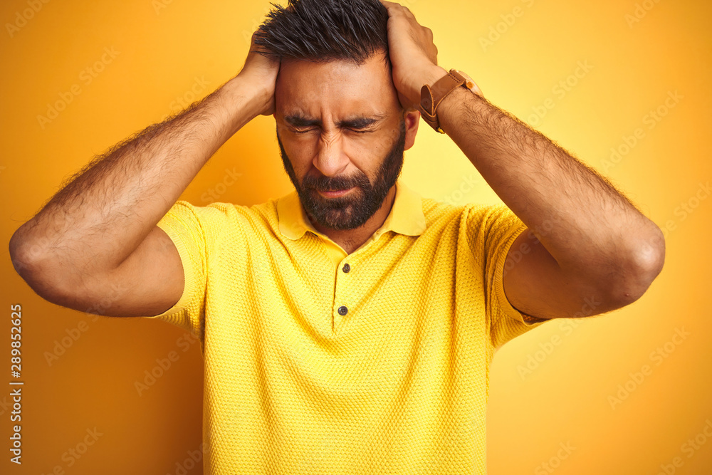 Young indian man wearing polo standing over isolated yellow background suffering from headache desperate and stressed because pain and migraine. Hands on head. Young indian man wearing polo standing over isolated yellow background suffering from headache desperate and stressed because pain and migraine. Hands on head.
