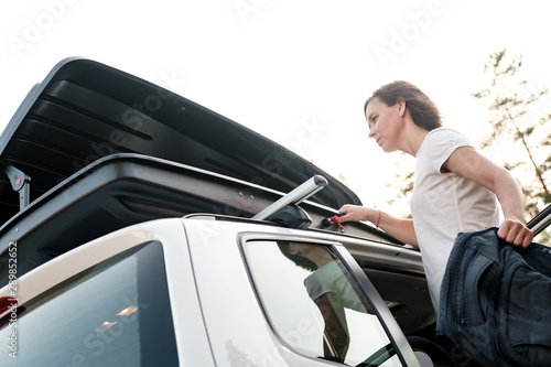 A woman puts things in the roof rack of a car or in a cargo box, before a family trip on vacation, against the sky and trees, on a summer evening.