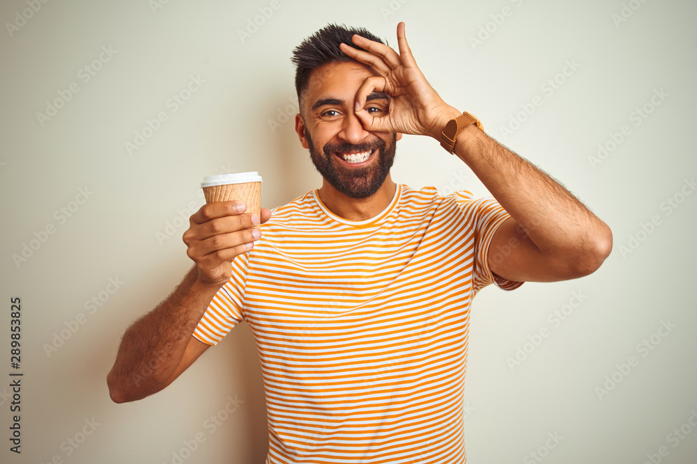 Young indian man drinking cup of coffee standing over isolated white background with happy face smiling doing ok sign with hand on eye looking through fingers Young indian man drinking cup of coffee standing over isolated white background with happy face smiling doing ok sign with hand on eye looking through fingers