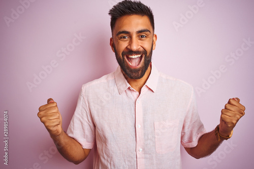 Young indian man wearing casual shirt standing over isolated pink background celebrating surprised and amazed for success with arms raised and open eyes. Winner concept.