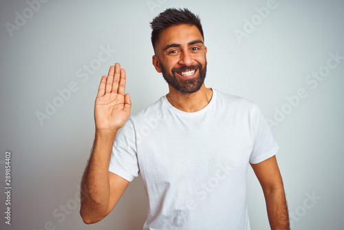 Young indian man wearing t-shirt standing over isolated white background Waiving saying hello happy and smiling, friendly welcome gesture photo