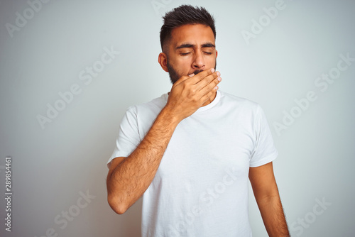 Young indian man wearing t-shirt standing over isolated white background bored yawning tired covering mouth with hand. Restless and sleepiness.