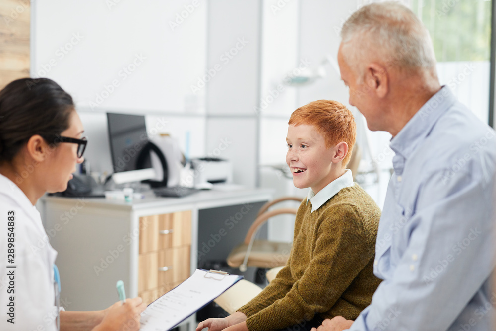 Side view portrait of smiling boy visiting doctor with father and looking at nurse filling patients form in waiting room, copy space Side view portrait of smiling boy visiting doctor with father and looking at nurse filling patients form in waiting room, copy space