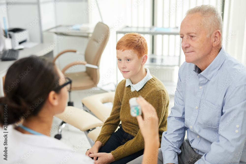 High angle portrait of cute red haired boy visiting doctor with father and looking at nurse holding pill bottle while prescribing vitamins, copy space High angle portrait of cute red haired boy visiting doctor with father and looking at nurse holding pill bottle while prescribing vitamins, copy space