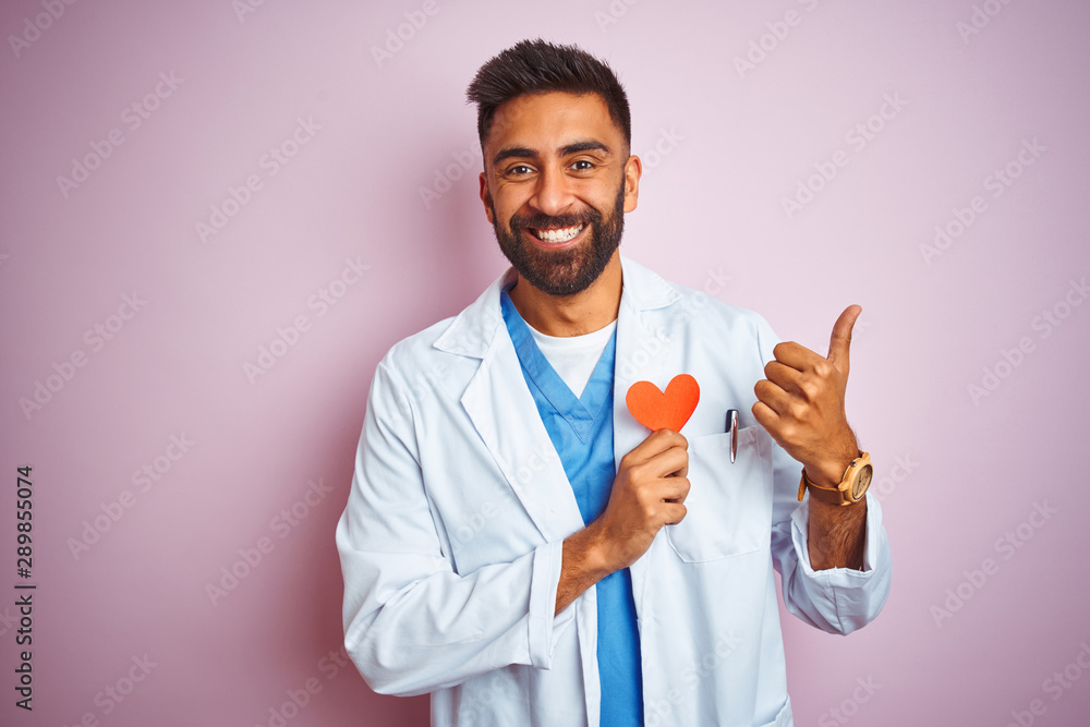 Young indian doctor man holding paper heart standing over isolated pink background happy with big smile doing ok sign, thumb up with fingers, excellent sign Young indian doctor man holding paper heart standing over isolated pink background happy with big smile doing ok sign, thumb up with fingers, excellent sign
