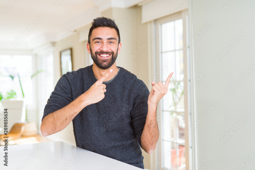 Handsome hispanic man wearing casual sweater at home smiling and looking at the camera pointing with two hands and fingers to the side.