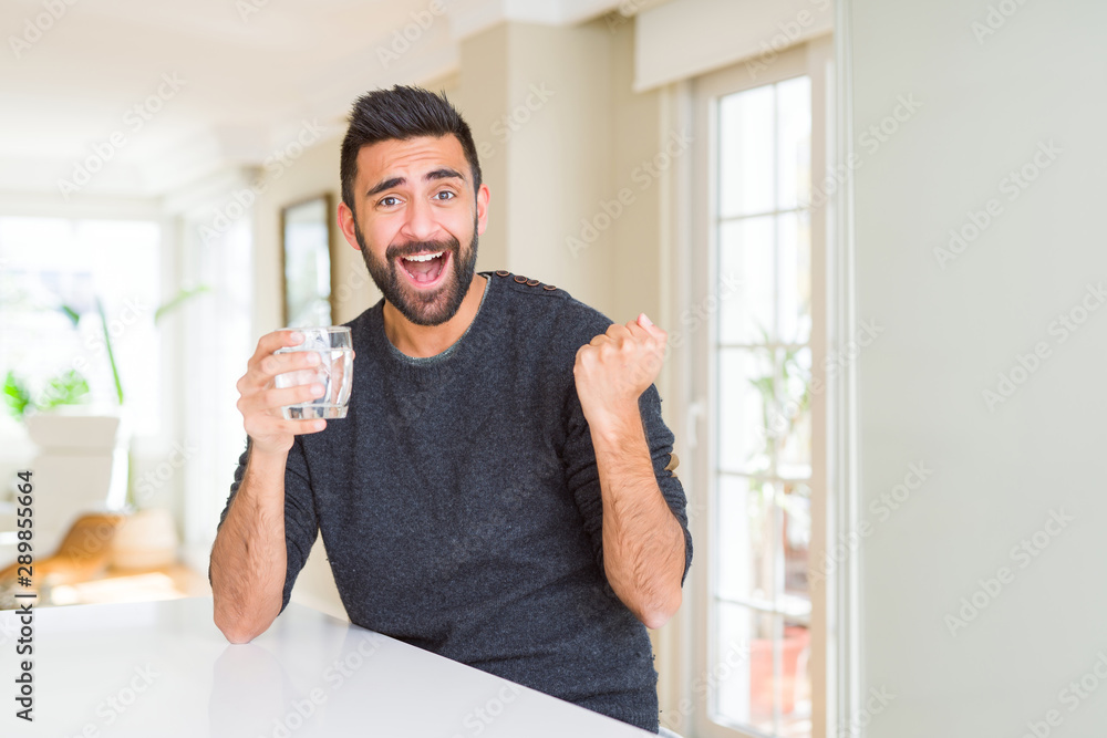 Handsome hispanic man drinking a fresh glass of water screaming proud and celebrating victory and success very excited, cheering emotion Handsome hispanic man drinking a fresh glass of water screaming proud and celebrating victory and success very excited, cheering emotion