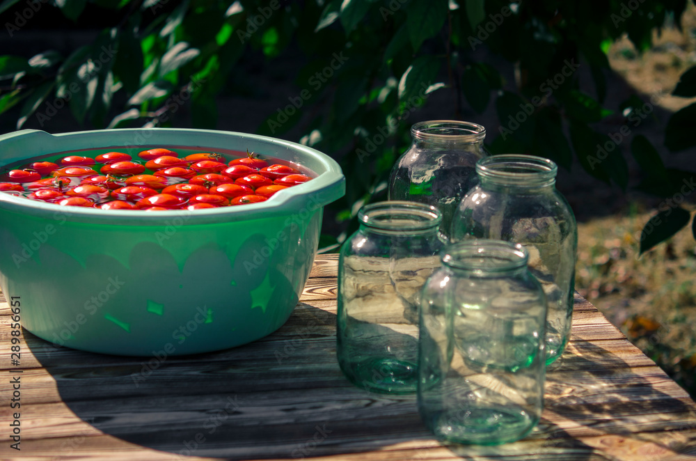 A fresh crop of red tomatoes washed in clear water, floats in a round plastic bowl, which stands on a wooden table, next to glass jars in the garden A fresh crop of red tomatoes washed in clear water, floats in a round plastic bowl, which stands on a wooden table, next to glass jars in the garden