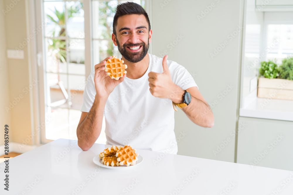 Handsome hispanic man eating sweet belgian waffle pastry happy with big smile doing ok sign, thumb up with fingers, excellent sign Handsome hispanic man eating sweet belgian waffle pastry happy with big smile doing ok sign, thumb up with fingers, excellent sign