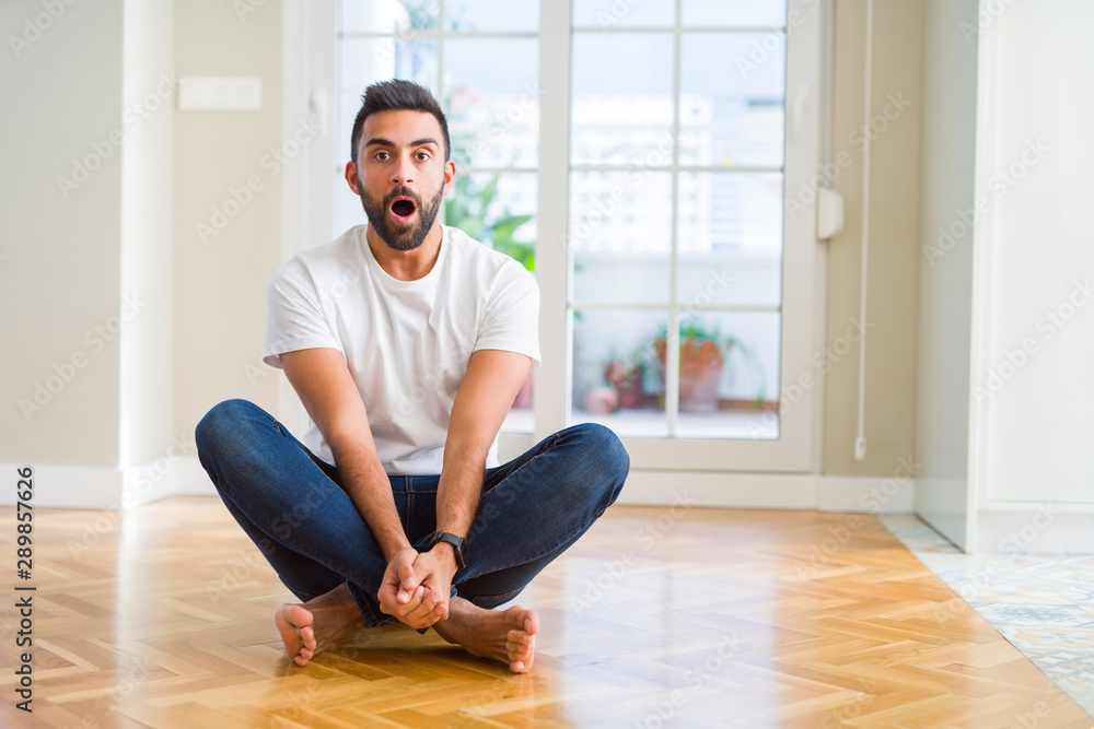 Handsome hispanic man wearing casual t-shirt sitting on the floor at home afraid and shocked with surprise expression, fear and excited face. Handsome hispanic man wearing casual t-shirt sitting on the floor at home afraid and shocked with surprise expression, fear and excited face.
