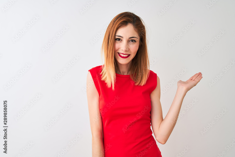 Redhead businesswoman wearing elegant red dress standing over isolated white background smiling cheerful presenting and pointing with palm of hand looking at the camera. Redhead businesswoman wearing elegant red dress standing over isolated white background smiling cheerful presenting and pointing with palm of hand looking at the camera.