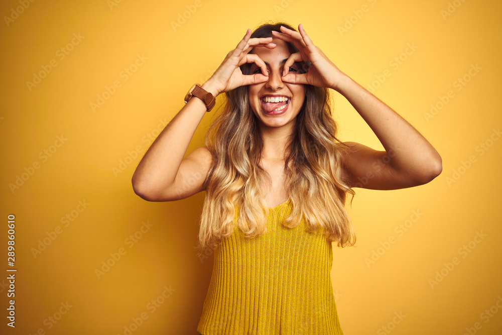 Young beautiful woman wearing t-shirt over yellow isolated background doing ok gesture like binoculars sticking tongue out, eyes looking through fingers. Crazy expression. Young beautiful woman wearing t-shirt over yellow isolated background doing ok gesture like binoculars sticking tongue out, eyes looking through fingers. Crazy expression.