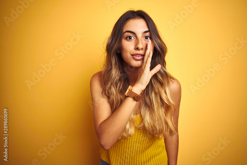 Young beautiful woman wearing t-shirt over yellow isolated background hand on mouth telling secret rumor, whispering malicious talk conversation photo