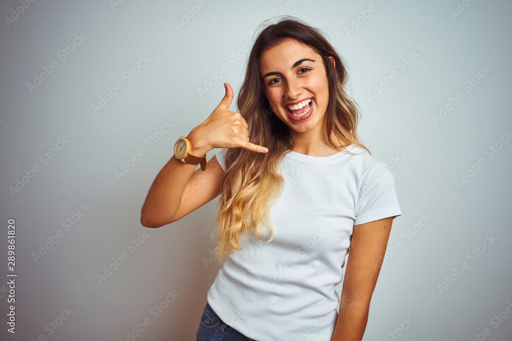 Young beautiful woman wearing casual white t-shirt over isolated background smiling doing phone gesture with hand and fingers like talking on the telephone. Communicating concepts. Young beautiful woman wearing casual white t-shirt over isolated background smiling doing phone gesture with hand and fingers like talking on the telephone. Communicating concepts.