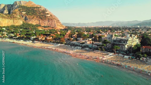 Palermo, SICILY, Italy - August 2019: Huge beach on the ocean. People swim and sunbathe. Against the background of a city with a lot of trees and plants. Rocky Mountains. Symmetric umbrellas on the