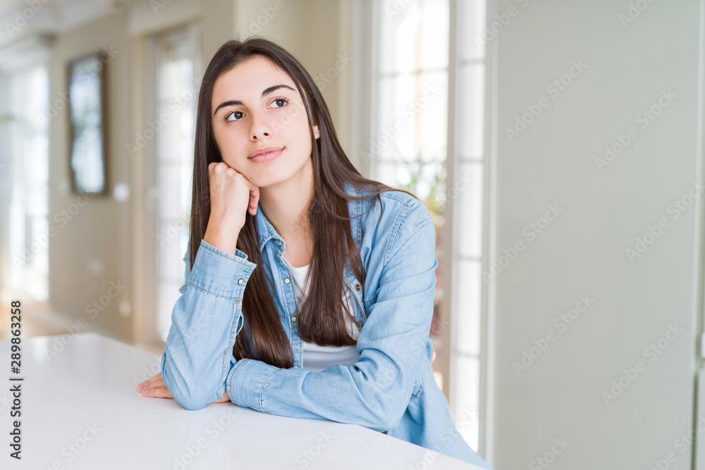 Beautiful young woman sitting on white table at home with hand on chin thinking about question, pensive expression. Smiling with thoughtful face. Doubt concept. Beautiful young woman sitting on white table at home with hand on chin thinking about question, pensive expression. Smiling with thoughtful face. Doubt concept.