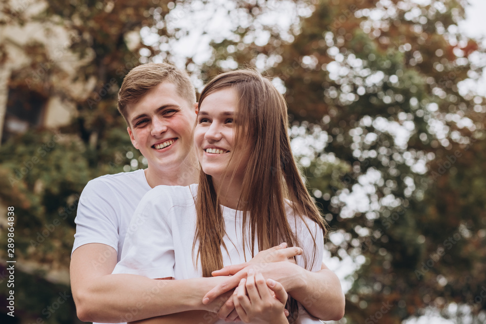Fototapeta premium Happy young couple walks the streets of the city and hold hands. guy and girl in white t-shirts and jeans outdoors. Teenagers cuddling against the backdrop of an autumn tree. Couple close-up portrait