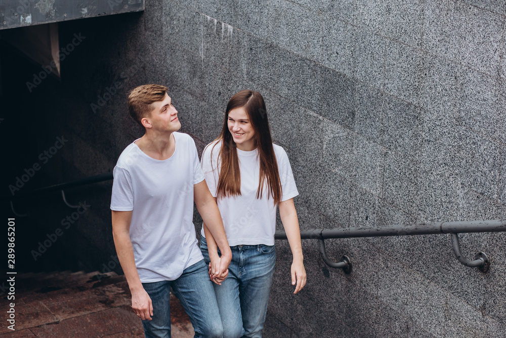Happy young couple walks the streets of the city and hold hands. guy and girl in white t-shirts and jeans outdoors. Teenagers hug on the background of the urban landscape. Couple close up portrait.