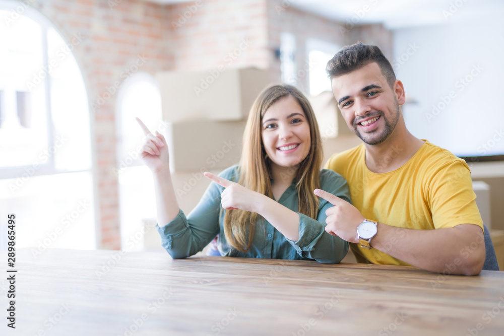 Young couple sitting on the table movinto to new home with carboard boxes behind them smiling and looking at the camera pointing with two hands and fingers to the side. Young couple sitting on the table movinto to new home with carboard boxes behind them smiling and looking at the camera pointing with two hands and fingers to the side.