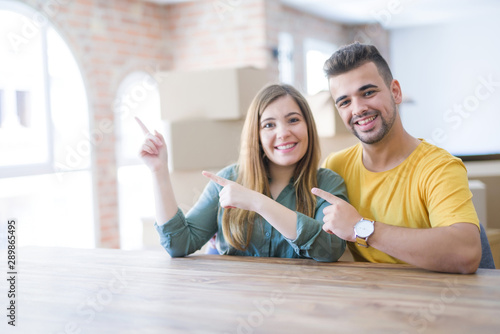 Young couple sitting on the table movinto to new home with carboard boxes behind them smiling and looking at the camera pointing with two hands and fingers to the side.