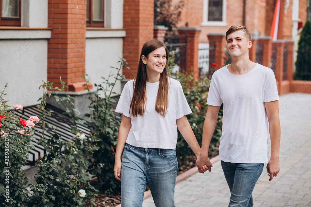 Happy young couple walks the streets of the city and hold hands. guy and girl in white t-shirts and jeans outdoors. Teenagers are walking in the autumn city on the background of a brick fence. Happy young couple walks the streets of the city and hold hands. guy and girl in white t-shirts and jeans outdoors. Teenagers are walking in the autumn city on the background of a brick fence.