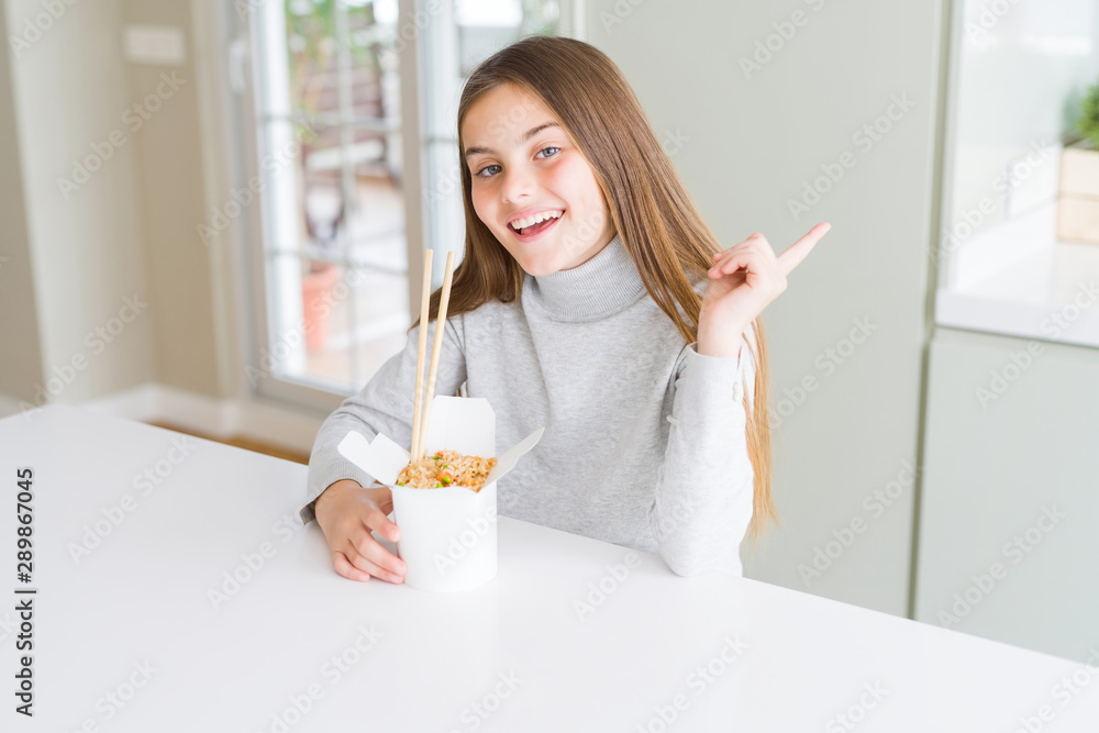 Beautiful young girl kid eating asian rice in delivery box very happy pointing with hand and finger to the side Beautiful young girl kid eating asian rice in delivery box very happy pointing with hand and finger to the side