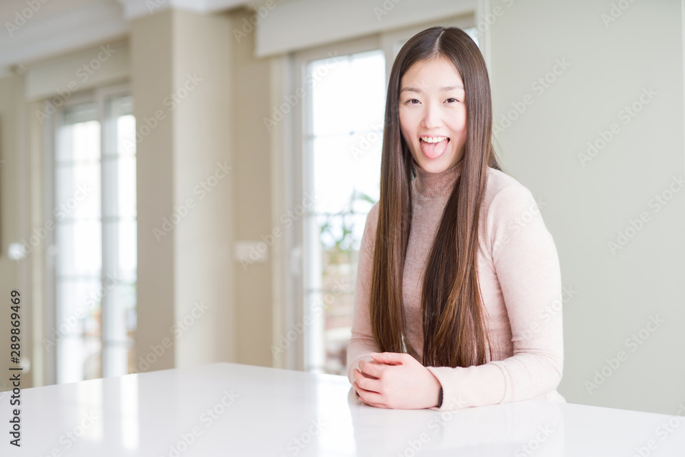 Beautiful Asian woman wearing casual sweater on white table sticking tongue out happy with funny expression. Emotion concept. Beautiful Asian woman wearing casual sweater on white table sticking tongue out happy with funny expression. Emotion concept.