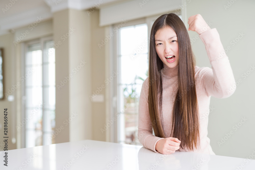Beautiful Asian woman wearing casual sweater on white table angry and mad raising fist frustrated and furious while shouting with anger. Rage and aggressive concept. Beautiful Asian woman wearing casual sweater on white table angry and mad raising fist frustrated and furious while shouting with anger. Rage and aggressive concept.