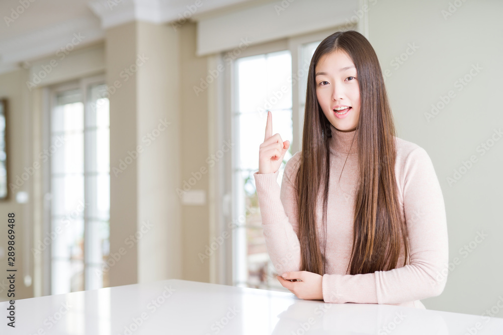 Beautiful Asian woman wearing casual sweater on white table pointing finger up with successful idea. Exited and happy. Number one. Beautiful Asian woman wearing casual sweater on white table pointing finger up with successful idea. Exited and happy. Number one.